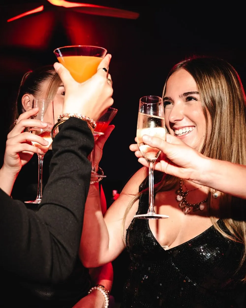Des amies trinquent, cocktail et champagne levés, une jeune femme rit aux éclats